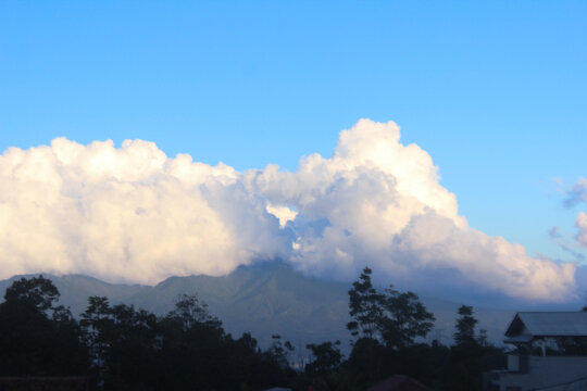A picturesque landscape featuring a mountain range partially obscured by large, fluffy cumulus clouds under a clear blue sky.