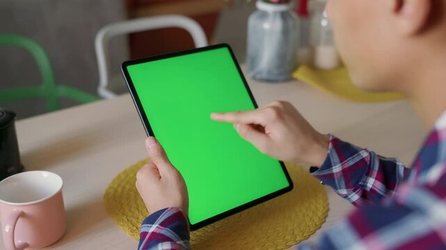 young man tapping tablet with green screen at cozy table, vertical view. using app, confirming action or starting video in relaxed home or cafe setting