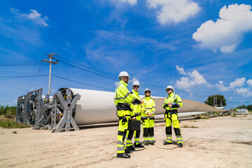Engineers in high visibility gear assess a large wind turbine blade at a construction site under a bright blue sky, representing the implementation of renewable energy technology for sustainable.