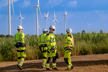 A team of engineers in high visibility safety gear stand together at a wind farm, conducting...
