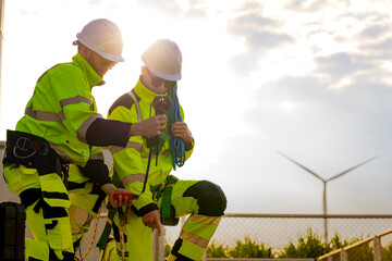 Two engineers wearing high visibility safety jackets and helmets measure wind speed with an...