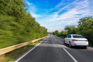 A white sedan drives swiftly down a forest-lined highway under a bright blue sky, with motion blur...