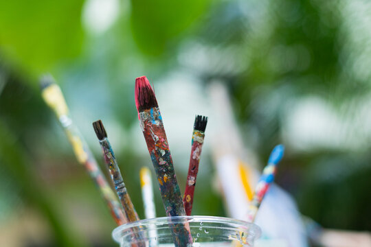 Selective focus with copy space of colorful paint brushes in a cup, splattered with paint, against a lush, blurred backdrop for creativity, art supplies, and artistic expression in nature outdoor.