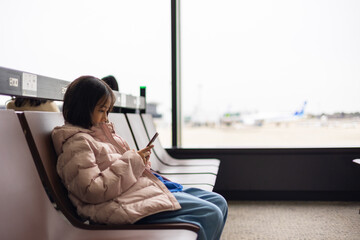 Adorable kid traveler in a winter jacket waits alone at the airport, engrossed in a mobile device. The blurred airplane view highlights solitude, digital connectivity, and modern travel habits.