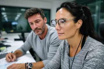 A focused woman in glasses collaborates with a serious Caucasian man in a modern office, both engaged in a discussion while analyzing documents
