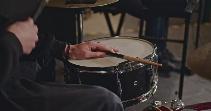 Close-up of a drummer's hands playing a snare drum with drumsticks in a dimly lit rehearsal space.