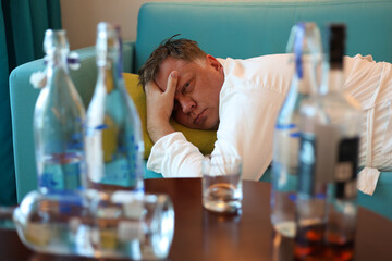 A middle aged man rests on a sofa near a table filled with bottles of alcohol, tired and sore after drinking too much.