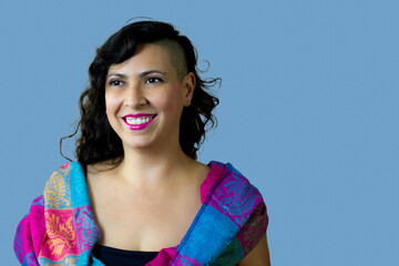 Confident woman with asymmetrical haircut and bright pink lipstick smiling and looking away. She wears colorful scarf and half-shaved hairstyle. Copy space on blue background.