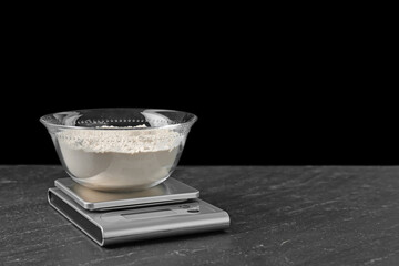 Electronic kitchen scale with bowl of flour on dark textured table against black background. Space for text