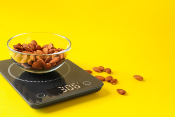 Electronic kitchen scale with bowl of almond nuts on yellow background, closeup. Space for text