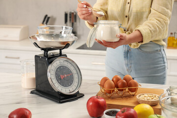Woman holding jar of flour and spoon near mechanical kitchen scale at white marble table indoors,...