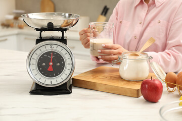 Woman with measuring cup of milk near mechanical kitchen scale at white marble table indoors,...