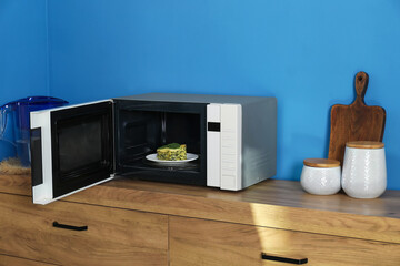 Open microwave oven with piece of lasagna and kitchen utensils on wooden cabinet indoors