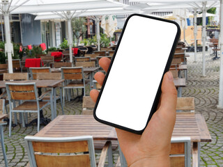 Smartphone with transparent screen in human hands. In the background, a garden restaurant with chairs, tables and parasols.