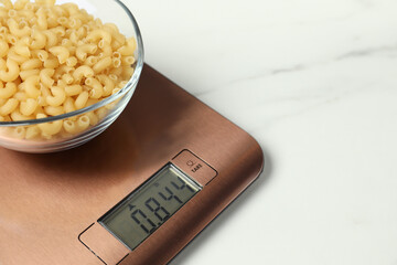 Modern electronic kitchen scale with bowl of raw pasta on white marble table, closeup. Space for...