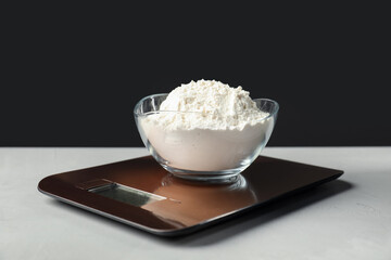 Electronic kitchen scale with bowl of flour on grey textured table against black background, closeup