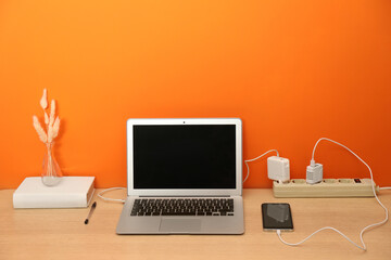 USB adapters with cables plugged into power strip charging laptop and smartphone on wooden table near orange wall