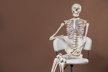 Artificial human skeleton model sitting on chair against light brown background, space for text