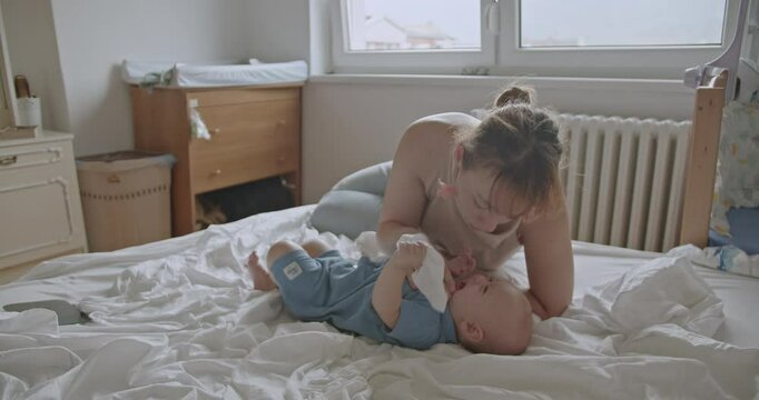 A mother plays with her baby boy on a bed in a softly lit room. They share smiles, laughs, and tender moments together.