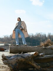 Naklejka premium Young woman stands on log by lake against background of blue sky with clouds.