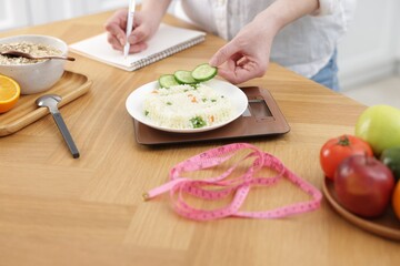 Woman weighting rice with vegetables and writing in notebook at wooden table, closeup