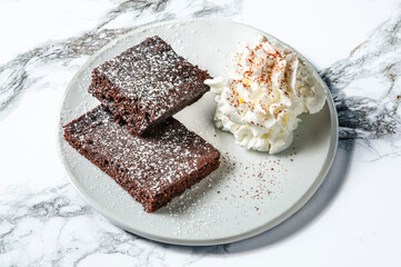 Two chocolate brownies dusted with powdered sugar, paired with whipped cream on a white plate. Elegant marble background adds a luxurious touch, emphasizing the dessert's rich brown tones and creamy w