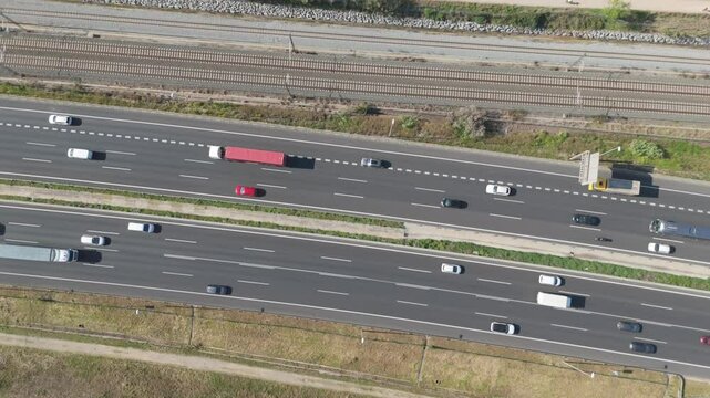 Direct top-down aerial view of highway traffic, including cars and a yellow truck, driving alongside parallel railway tracks. Strong lines and daytime shadows.

