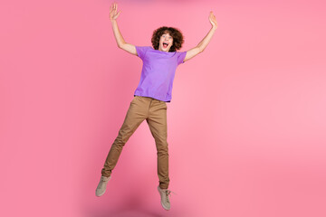 Young man with curly brown hair in purple t-shirt joyfully jumps against solid pink background,...