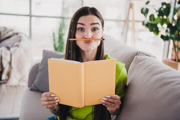 Cheerful young woman playfully balances a pencil while reading a book on a cozy couch in a bright and sunny home interior