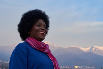 Joyful Afroamerican woman with vibrant scarf smiling in front of snowy mountain range at dusk
