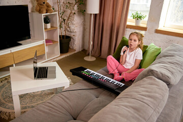 Little girl sitting on sofa with microphone singing while watching online lesson on laptop, learning music performance online. Concept of childhood, online education, remote studying