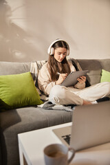 Obraz premium Focused teen girl sitting cross-legged on sofa with headphones, writing on digital tablet during remote learning session in modern room interior. Concept of education, homeschooling, studying