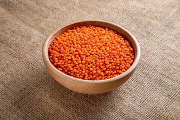 A wooden bowl with red lentils grains on a burlap. Top view of dried orange lentils inside the bowl.