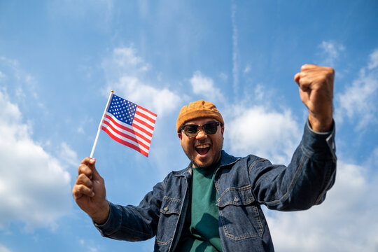 Portrait of cheerful young man looking forward while standing proudly with holding USA flag over his head against wind and blue sky, happy american holding the United states flag celebration