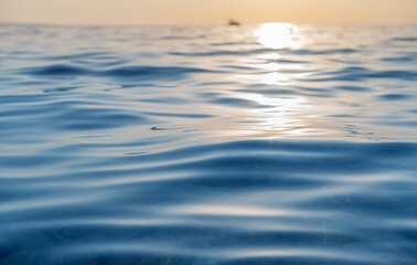 A serene seascape captures gentle ocean ripples reflecting the golden glow of the setting sun, with a small boat silhouetted on the distant horizon.