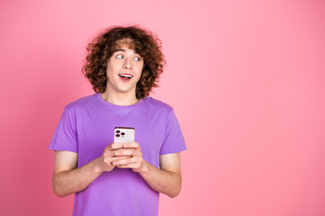 Cheerful young man with curly brown hair holding a smartphone, dressed in a purple t-shirt,...
