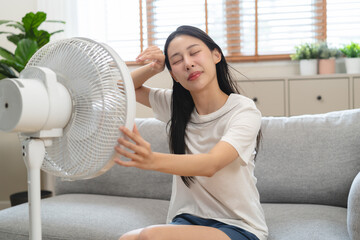 Suffer summer heat wave stroke, hot weather, tired asian young woman sitting on couch in living room without air-conditioner, using electric fan, wave fan to cool for ventilation when high temperature