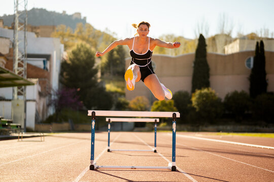 A pretty girl in her 20s and 30s in sportswear trains outdoor athletics.Front shot of girl jumping over a fence. Concept of women jumping hurdles.