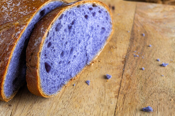 Freshly baked bread with a vibrant purple interior, cooked in blue Thai ancha tea, rests on a rustic wooden cutting board with a slice cut off to reveal its soft, crumbly texture, with copy space