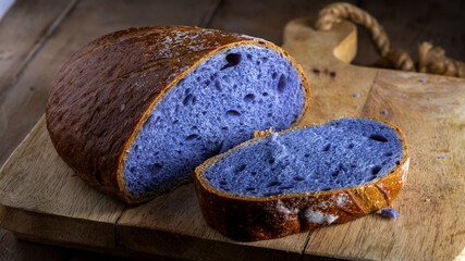 Freshly baked bread with a vibrant purple interior, cooked in blue Thai ancha tea, rests on a rustic wooden cutting board with a slice cut off to reveal its soft, crumbly texture.