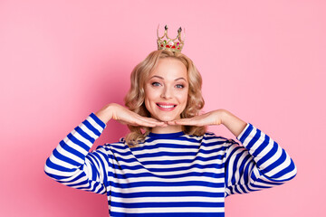 Cheerful young woman in a striped pullover posing with a playful gesture and wearing a crown on a pink background