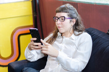 Fototapeta na wymiar Smiling young woman wearing glasses looking at cell phone doing mobile shopping relaxing on sofa in living room. Happy mature young woman using smartphone sitting on couch at home
