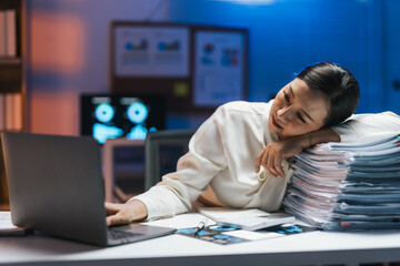 Tired doctor resting her head on a stack of documents while using a laptop, working late in the...