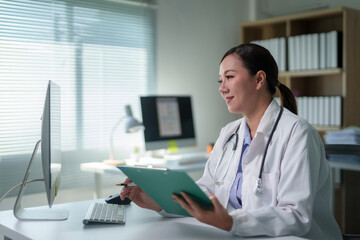 Asian female doctor reviewing medical records on a clipboard while working on a computer at her...