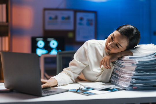 Tired doctor resting her head on a stack of documents while using a laptop, working late in the hospital