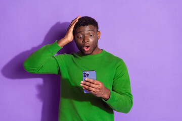 African American man in green sweatshirt looking surprised at smartphone against purple background,...