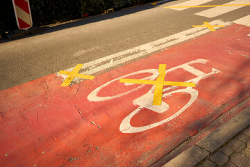 A red bike lane marked with yellow X tape, signaling closure or restriction. Urban textures, cracked asphalt, and morning light tell a silent story of change and interruption.