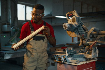 Serious carpenter in protective eyeglasses examining wooden detail in factory
