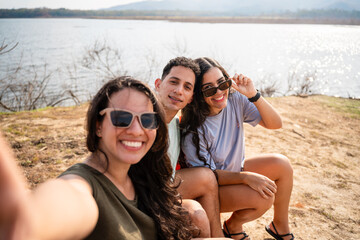 Fototapeta premium Latin american friends enjoying a summertime selfie by a scenic lake surrounded by nature and mountains