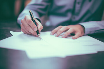 Naklejka premium Person signing a document with a pen in a modern office, focusing intently on the paper. Light streams in through a large window, casting shadows on the desk.